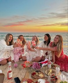 four women sitting on a wooden deck with food and drinks in front of the ocean