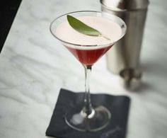 a drink in a martini glass with a green leaf on the rim next to a cocktail shaker