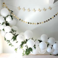 white balloons and garlands are hanging from the ceiling in an indoor space that is decorated with greenery