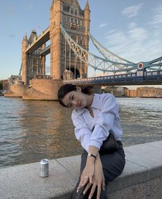 a woman sitting on the edge of a wall next to a body of water with a bridge in the background