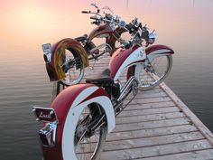 two motorcycles are parked on a dock near the water at sunset or sunrise, and one is red with white stripes