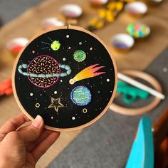 a person holding up a small embroidery project with space and stars on it in front of other crafting supplies