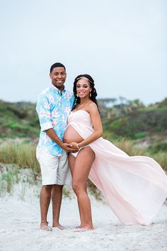 a pregnant woman in a pink dress poses with her husband on the beach for a photo