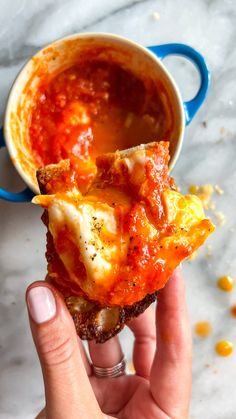 a hand holding up a piece of pizza with sauce and cheese on it in front of a cup