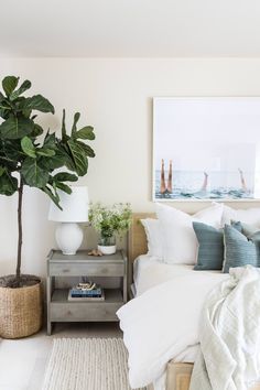 a bed with white sheets and pillows next to a potted plant on a nightstand