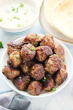 a white bowl filled with meatballs next to a plate of pita bread and dipping sauce