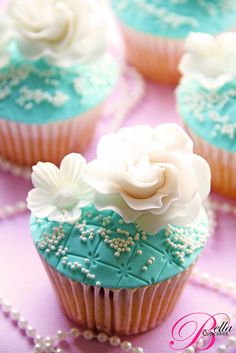 cupcakes decorated with white and blue frosting on a pink tablecloth next to pearls