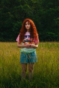 a woman with red hair standing in tall grass wearing a t - shirt and shorts