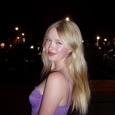 a woman with long blonde hair standing in front of a street at night wearing a purple dress