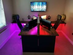 a desk with two monitors and a keyboard on it in a room that has purple lighting