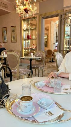 a woman sitting at a table with two cups of coffee in front of her and a chandelier hanging from the ceiling