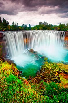 the waterfall is surrounded by lush green grass and trees, under a dark cloudy sky