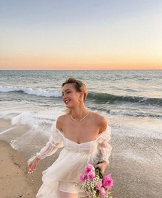 a woman is walking on the beach with flowers in her hand and smiling at the camera