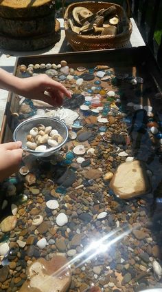 two hands reaching for rocks on top of a glass table covered in pebbles and shells