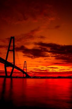 the sun is setting behind a bridge over water with buildings in the distance and red sky