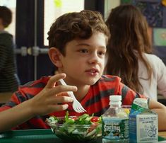 a young boy sitting at a table eating salad with a fork and bottle of water