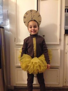 a young child wearing a tutu and hat standing in front of a door with a clock on it's face