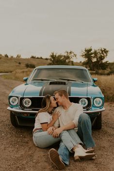 a man and woman sitting on the ground next to a blue mustang in front of a car