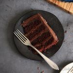 a slice of chocolate cake on a black plate with a fork and knife next to it