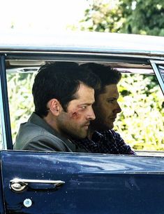 two men sitting in the back seat of a blue car with blood on their face