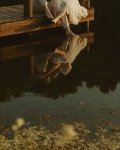 a man and woman sitting on a dock next to water with their reflection in the water