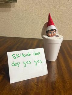 an elf is sitting in a cup next to a sign that says skibia dap do yes yes