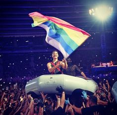 a man holding a rainbow flag in front of a crowd at a concert with his hands up