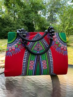 Stand out with this attractive large red dashiki African waxprint bag! This bag will make any outfit pop! This is a large-sized bag with a large carrying capacity and with a hard board shaping the entire purse, it is extra sturdy. There is a black lining, and interior zipper to securely close the bag. The straps are made from leather and wooden beads. The approximate dimensions of the bag are: 16.7"wide 13" high Straps: 6" For more African paintings, handbags, jewelry and fashion accessories, ba Traditional Red Rectangular Satchel, African Paintings, Top Handle Bags, Wooden Beads, Purses And Handbags, Top Handle, Top Handle Bag, Bathing Beauties, Accessory Gift