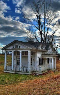 an old white house sitting in the middle of a field with trees and clouds above it