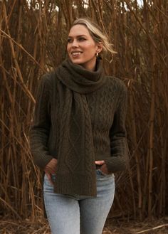 a woman standing in front of tall grass wearing a green cabled sweater and scarf