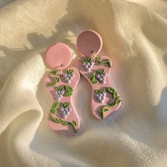 two pairs of pink earrings with green leaves and berries on them sitting on a white sheet