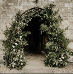 an arch covered in flowers and greenery next to a stone building with a door