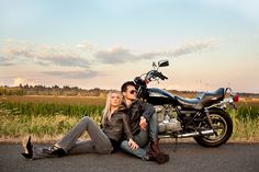 a man and woman sitting on the ground next to a motorcycle in front of a field