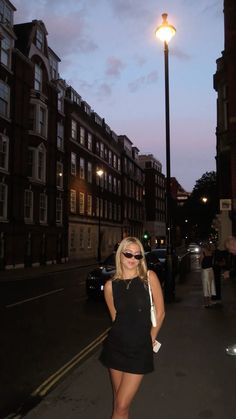 a woman standing on the side of a street next to tall buildings at night with her hands in her pockets