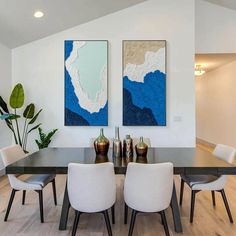 a dining room table with white chairs and two paintings on the wall above it, along with potted plants