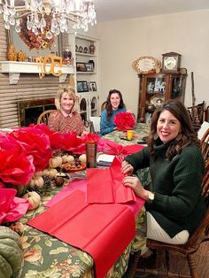 three women sitting at a table with red paper flowers on it and one woman standing in front of the table