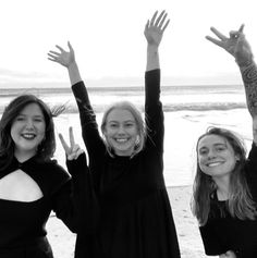 three women standing on the beach with their arms in the air and one holding her hands up