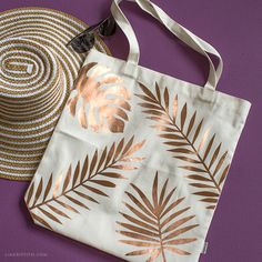 a white bag with gold leaves on it next to a straw hat and purple background