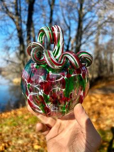 a hand holding up a glass candy cane in front of a lake and trees with leaves on the ground