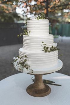 a white wedding cake sitting on top of a round table with greenery and lights in the background