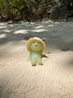 a small toy bear wearing a yellow hat on top of a sandy beach with trees in the background