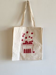 "This 100% cotton tote bag features a hand drawn basket full of cherries. This bag is good for the farmer's market, as a grocery tote bag, for school or every day use. It is printed here in dark red ink. We screen print these tote bags to order in our shop in Connecticut. Since these tote bags are each hand crafted and made to order, there may be slight variations in placement, but they are all beautiful! // FABRIC: 100% cotton // COLOR: Printed with red ink. Bag color is natural cotton (off-whi Natural Cotton Grocery Bag, Natural Cotton Grocery Bags, Reusable Cotton Bags For Grocery Shopping, Reusable Cotton Grocery Shopping Bags, Eco-friendly Cotton Bags For Farmers Market, Eco-friendly Red Cotton Canvas Bag, Organic Cotton Canvas Bag For Gift, Organic Cotton Bags For Gift, Organic Cotton Bag For Gift