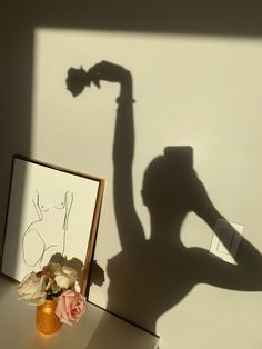 the shadow of a person holding a flower in front of a framed photograph and vase with flowers