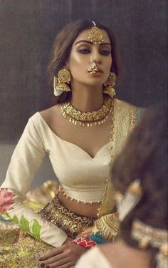 a woman in white dress with gold jewelry and nose ring sitting on a table next to another woman