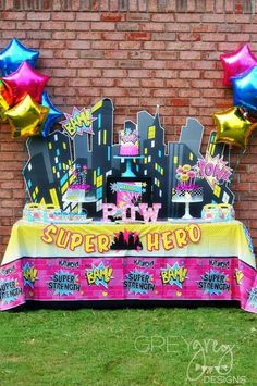 a table with balloons and decorations on it in front of a brick wall that says super hero