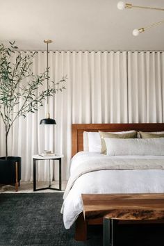 a bed with white sheets and pillows next to a tall plant in a room that has vertical blinds on the wall