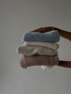 a person holding three folded sweaters in their hands, one is white and the other is blue