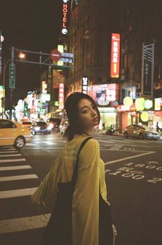 a woman standing in the middle of a street at night with cars and neon signs behind her