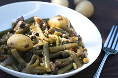 a white bowl filled with green beans and potatoes next to silverware on a table