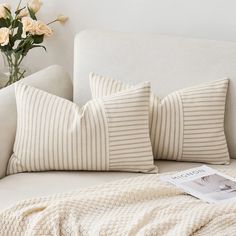 a white couch with two pillows and a magazine on it next to flowers in a vase